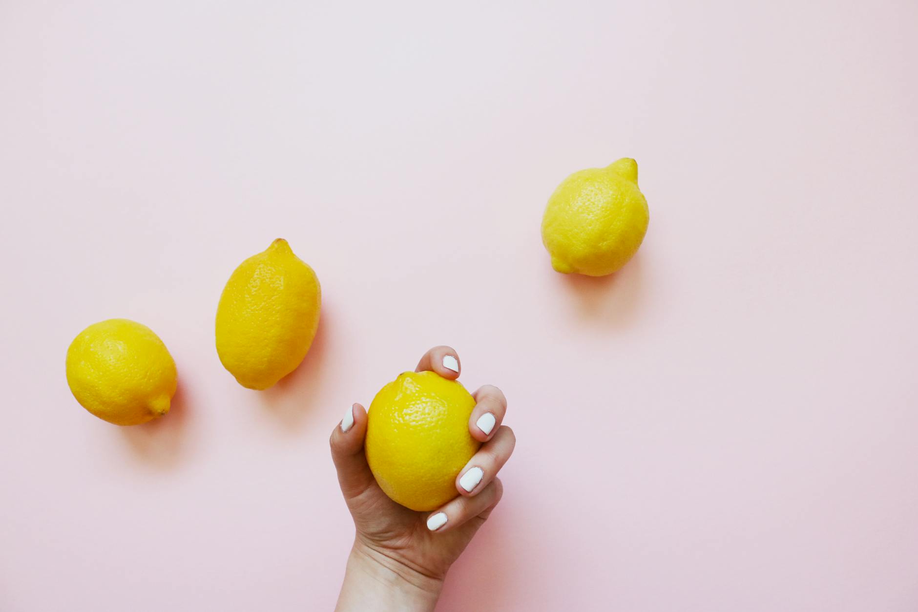 O mână care ține un lemon galben viu pe un fundal roz deschis, înconjurat de citrice proaspete