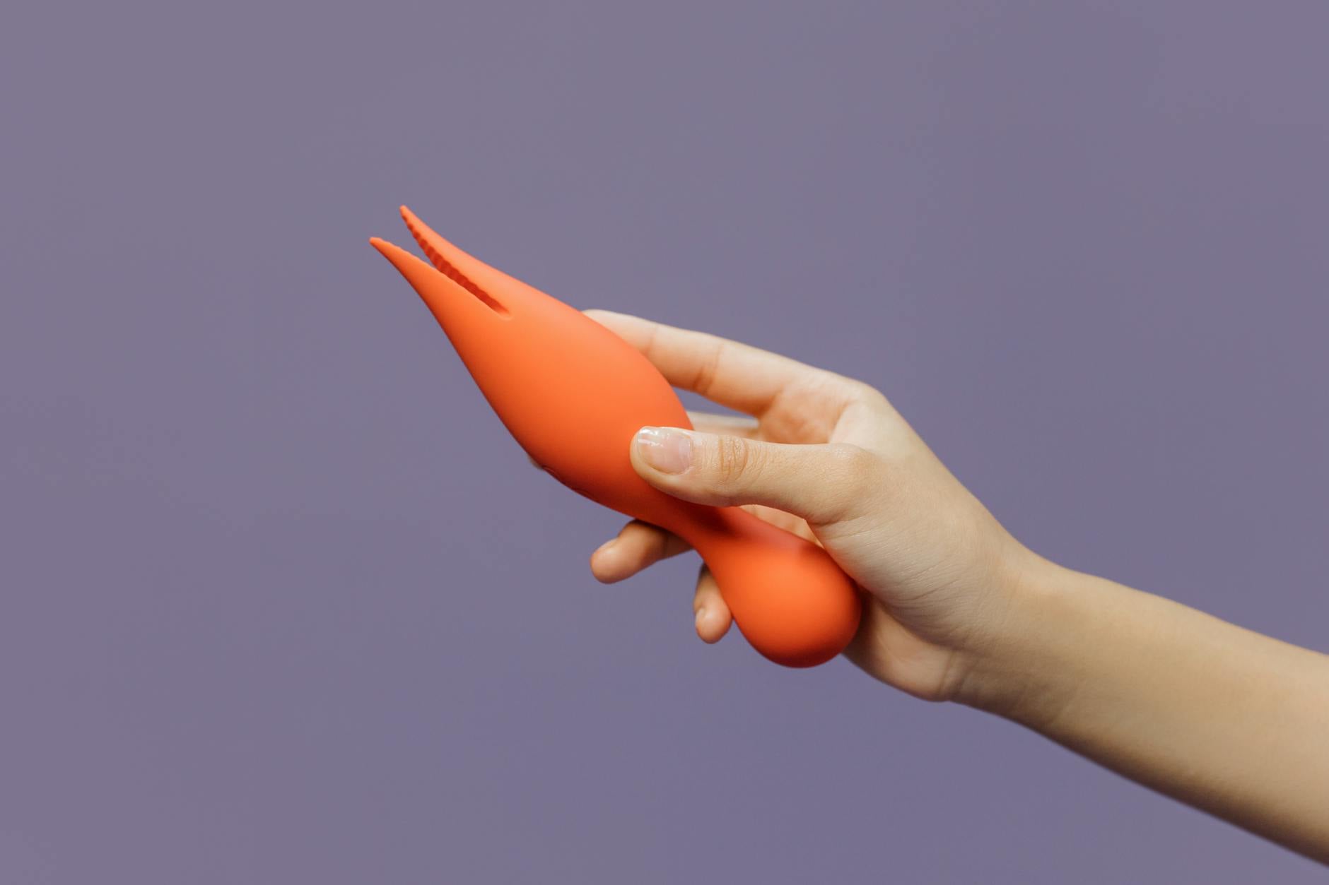 Mână ținând vibrator portocaliu pe fundal violet calmant, reprezentând revenirea sigură la plăcere